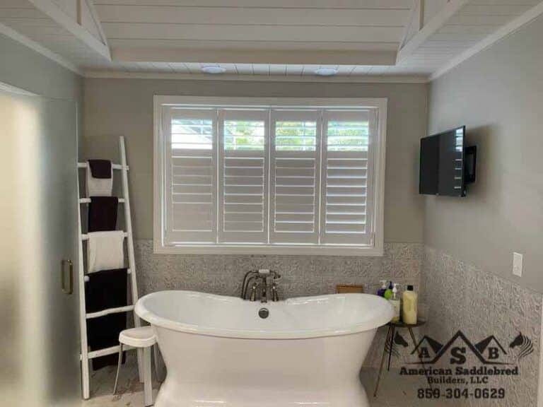 Freestanding bathtub positioned under a large window with plantation shutters